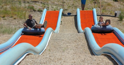 Two people on inflatable tubes slide down orange water slides on a sunny day, surrounded by gravel and grass.