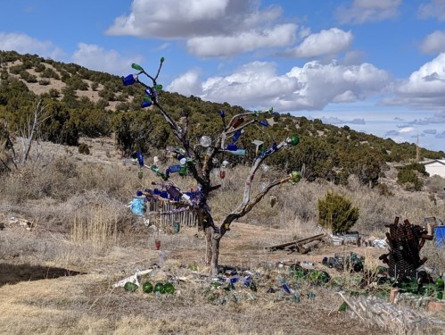 A whimsical tree decorated with colorful glass bottles, set against a backdrop of rolling hills and cloudy skies.