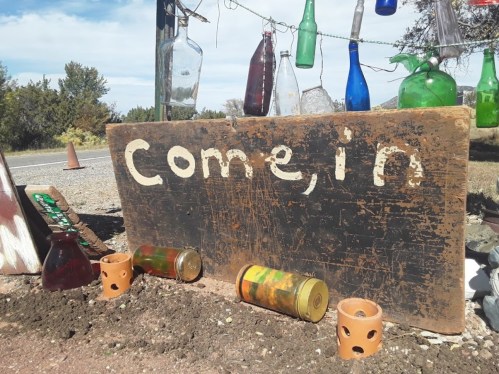 A weathered wooden sign reading "Come in" surrounded by colorful glass bottles and small clay pots.