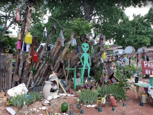 A quirky outdoor display featuring colorful bottles, sculptures, and a green alien figure among trees and rocks.