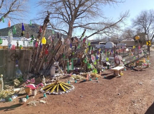 Colorful decorations and sculptures adorn a rustic outdoor space, surrounded by trees and a dirt path.