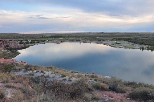 A serene landscape featuring a calm lake reflecting the sky, surrounded by grassy hills and distant structures.