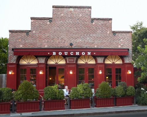 A charming brick restaurant with red accents and large windows, featuring outdoor seating and lush greenery.