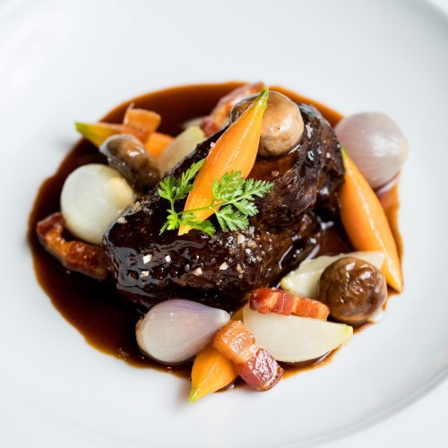 A beautifully plated dish featuring braised meat, carrots, mushrooms, and pearl onions in a rich sauce, garnished with herbs.