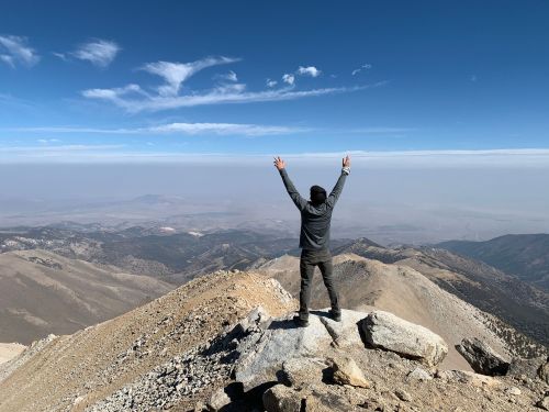 A person stands on a mountain peak, arms raised in triumph, with a vast landscape and blue sky in the background.