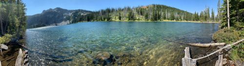 A serene lake surrounded by trees and mountains, with clear water reflecting the blue sky and sunlight.