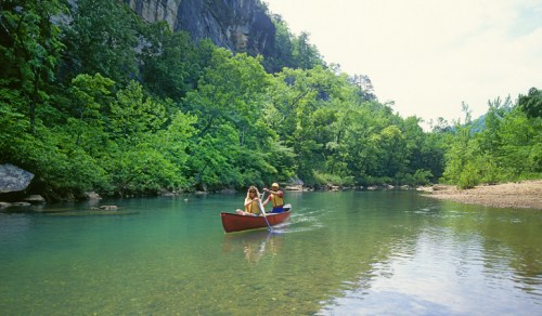 Two people paddle a red canoe on a calm river surrounded by lush green trees and rocky cliffs.