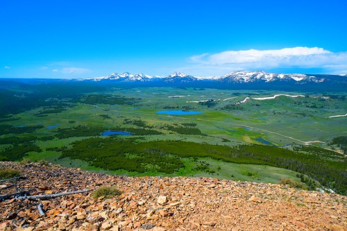 A panoramic view of lush green valleys, lakes, and snow-capped mountains under a clear blue sky.