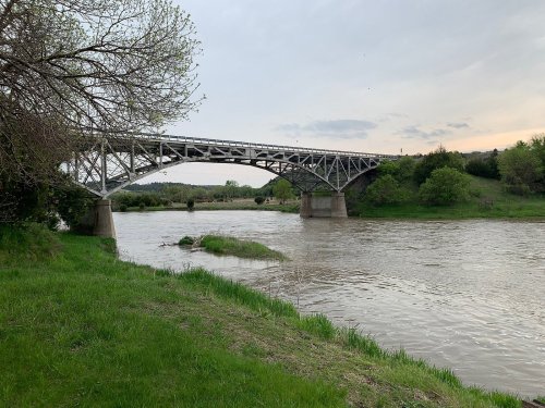 A metal bridge spans a river, surrounded by green grass and trees under a cloudy sky.