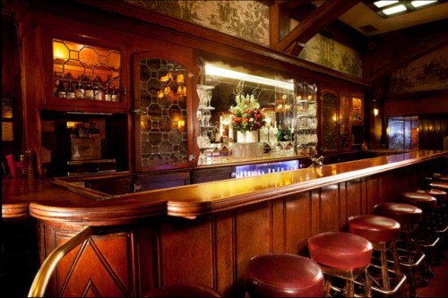 A warm, inviting bar with wooden paneling, a polished counter, and elegant floral arrangements behind the glass shelves.