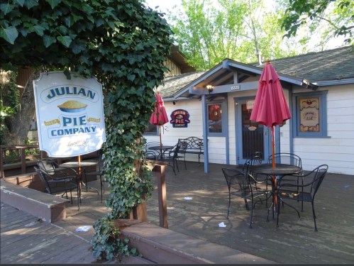 Exterior of Julian Pie Company with a sign, outdoor seating, and red umbrellas under green foliage.