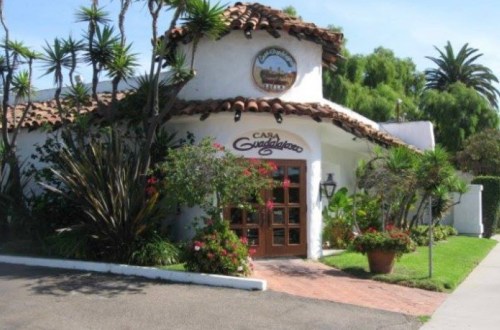 A charming white building with a tiled roof, surrounded by lush greenery and colorful flowers, featuring a sign that reads "Casa Guadalajara."
