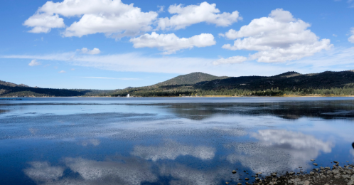 A serene lake reflecting clouds and mountains under a clear blue sky, surrounded by lush greenery.