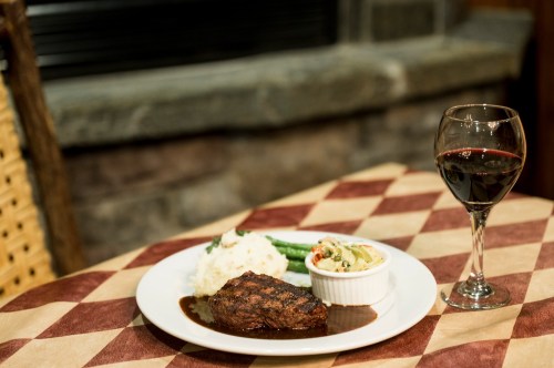 A plate with grilled steak, mashed potatoes, green beans, and a side dish, accompanied by a glass of red wine.