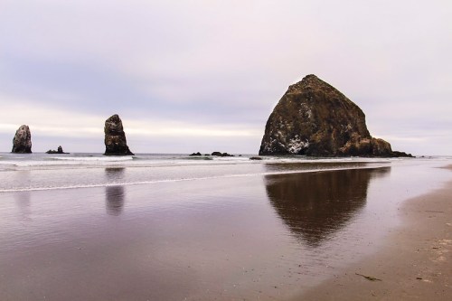 A serene beach scene with three large rock formations reflected in the calm water under a cloudy sky.