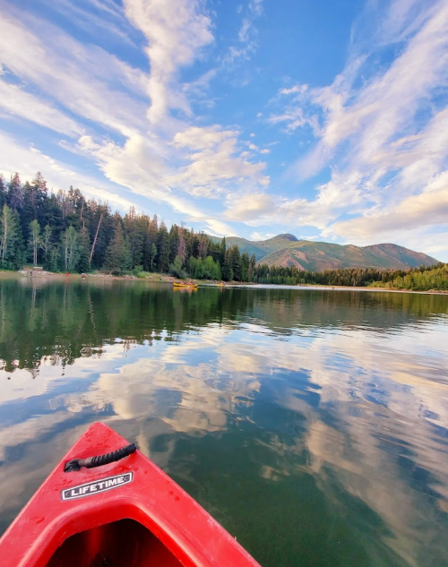 A red kayak on calm water reflects trees and clouds under a blue sky, with mountains in the background.