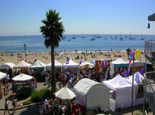 A vibrant beach scene with tents, people, and sailboats in the background under a clear blue sky.