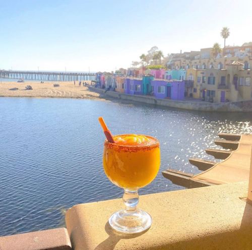 A vibrant mango drink in a glass sits on a ledge, with colorful buildings and a beach in the background.