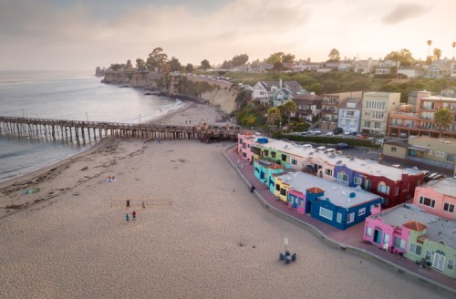 Aerial view of colorful beach houses along the shore, with a pier and cliffs in the background under a cloudy sky.