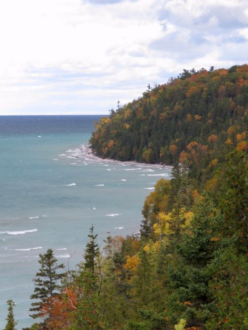 A scenic view of a coastline with vibrant autumn foliage and waves gently lapping at the shore under a cloudy sky.