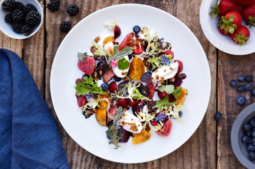 A vibrant salad with mixed greens, berries, edible flowers, and dollops of creamy dressing on a white plate.