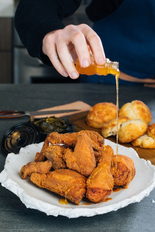 A hand drizzles honey over a plate of crispy fried chicken, with biscuits and greens in the background.