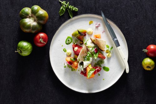 A beautifully plated dish featuring colorful tomatoes, herbs, and a slice of bread on a textured black background.