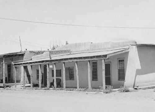 Black and white photo of a simple, old building with a flat roof and wooden posts, set on a quiet street.