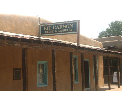 Sign for Kit Carson Home & Museum on a adobe-style building with wooden beams and blue window frames.