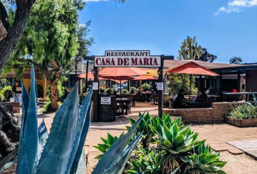 Outdoor view of Casa de Maria restaurant with patio seating, umbrellas, and surrounding greenery under a blue sky.
