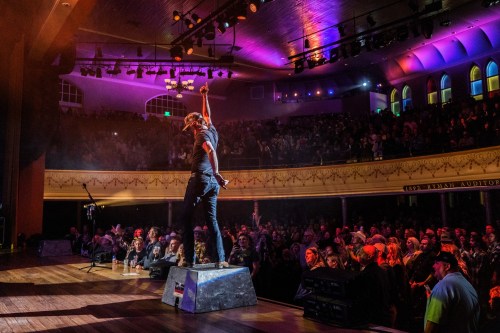 A performer stands on stage with one arm raised, energizing a large crowd in a vibrant, illuminated auditorium.