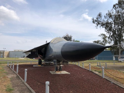A black military jet displayed outdoors on a gravel base, surrounded by trees and a blue sky.