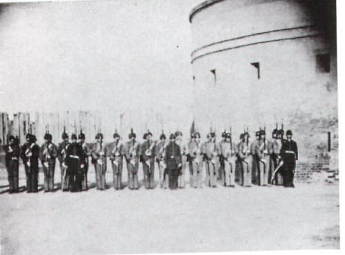 A historical black-and-white photo of soldiers in uniform standing in a line near a fortification.
