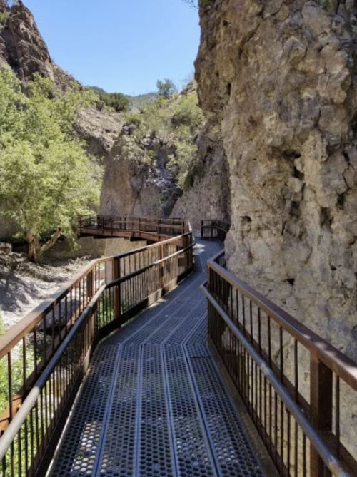 A winding metal walkway through a rocky canyon, surrounded by greenery and steep cliffs under a clear blue sky.