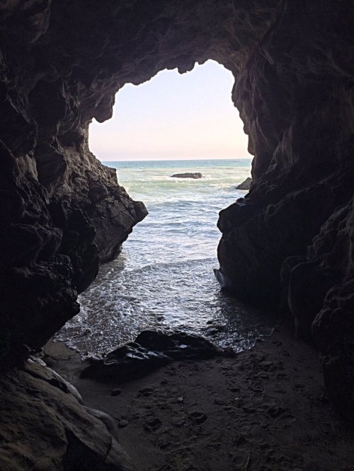 View from inside a cave looking out at the ocean, with waves gently lapping at the shore under a clear sky.