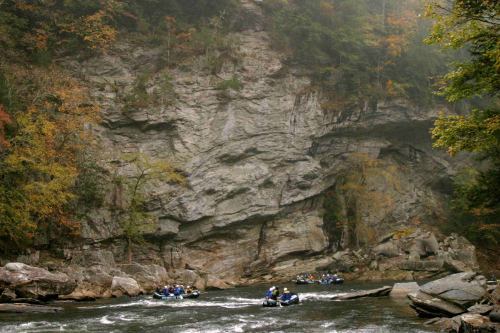 Rafting boats navigate a river surrounded by rocky cliffs and autumn foliage in a misty landscape.