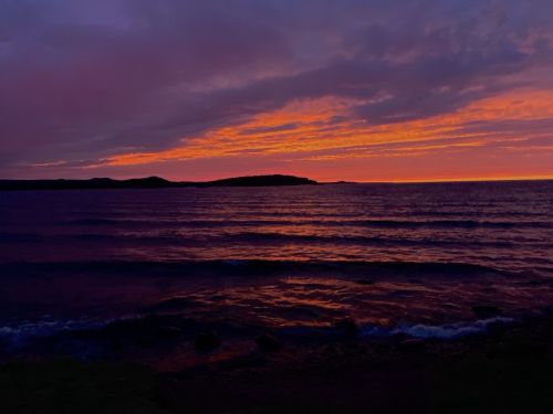 A vibrant sunset over calm waters, with purple and orange hues reflecting on the surface and silhouettes of distant hills.