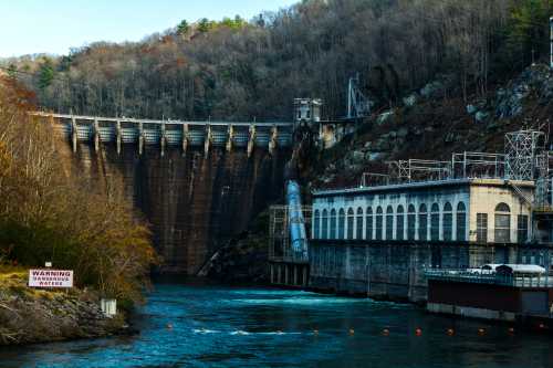 A large dam with a power station beside a river, surrounded by trees and rocky terrain, with a warning sign nearby.