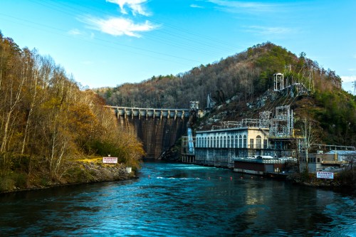 A large dam with a hydroelectric power station beside a river, surrounded by trees and a clear blue sky.