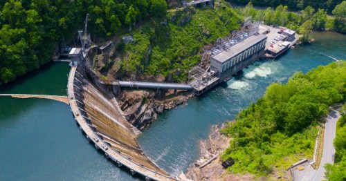 Aerial view of a dam surrounded by greenery, with water flowing over the spillway and a power station nearby.