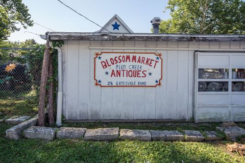 Sign for Blossom Market Antiques on a white building, surrounded by greenery and stone pathway.