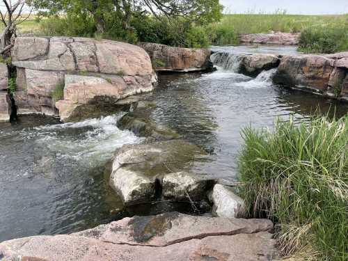 A serene stream flows over rocky formations, surrounded by lush greenery and gentle hills in the background.