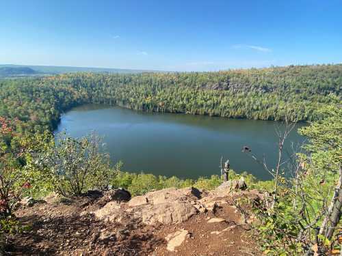 A serene view of a calm lake surrounded by lush green trees and rocky cliffs under a clear blue sky.