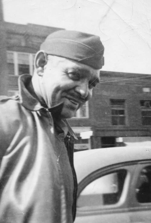 A smiling man in a military cap and leather jacket stands beside a vintage car, with a city street in the background.