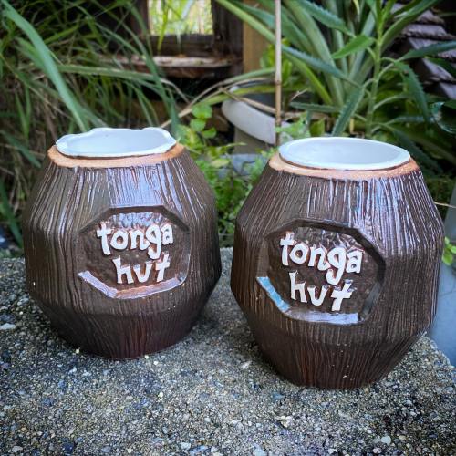 Two brown ceramic mugs with "Tonga Hut" embossed on them, placed on a textured surface with greenery in the background.