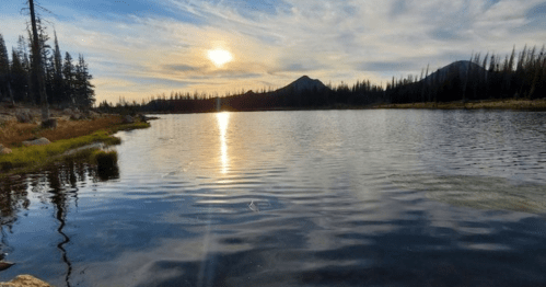 A serene lake at sunset, reflecting the sky and mountains, surrounded by trees and rocky shores.