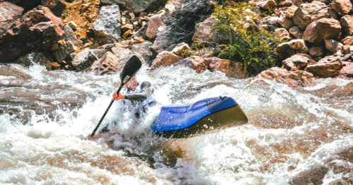A kayaker navigates through turbulent whitewater rapids, surrounded by rocky terrain and splashing water.