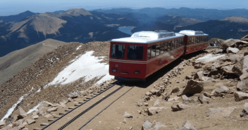 A red train travels along a rocky mountain track with scenic views of distant peaks and a lake below.