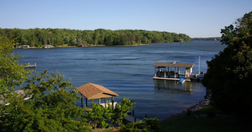A serene lake view with a dock and a boathouse, surrounded by lush greenery under a clear blue sky.