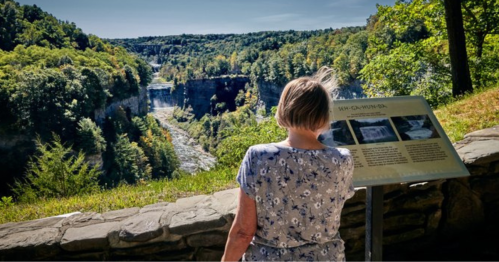 A person stands by a sign, overlooking a scenic view of a river and lush green landscape.